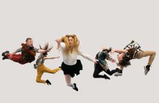 Five dancers in brightly coloured costumed leap in the air at different angles against a plain white backdrop.