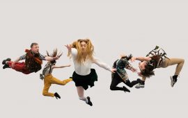 Five dancers in brightly coloured costumed leap in the air at different angles against a plain white backdrop.