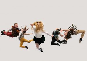 Five dancers in brightly coloured costumed leap in the air at different angles against a plain white backdrop.