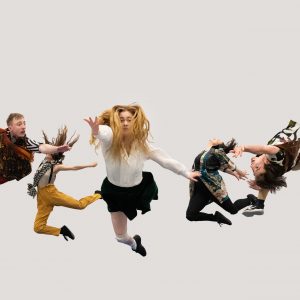 Five dancers in brightly coloured costumed leap in the air at different angles against a plain white backdrop.