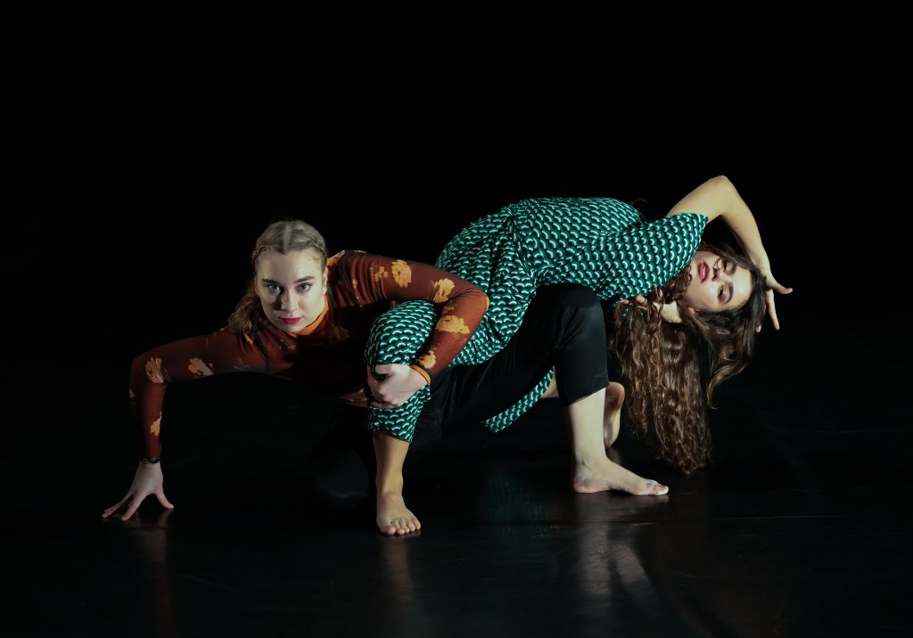 Two female Caucasian dancers in patterned clothing are are crouching intertwined with one another in front of a black background.