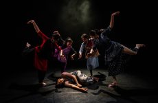 A group of four dancers pose around a fifth, who is lying on the floor as if dead or injured.