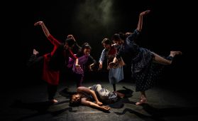 A group of four dancers pose around a fifth, who is lying on the floor as if dead or injured.