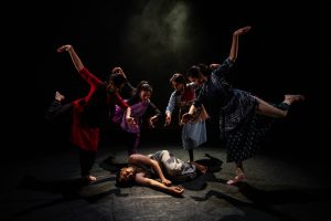 A group of four dancers pose around a fifth, who is lying on the floor as if dead or injured.
