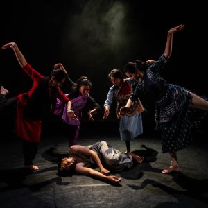 A group of four dancers pose around a fifth, who is lying on the floor as if dead or injured.
