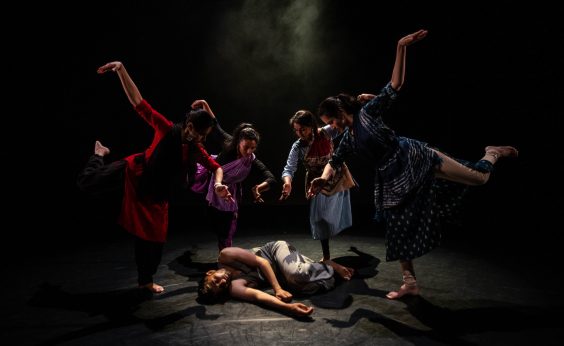 A group of four dancers pose around a fifth, who is lying on the floor as if dead or injured.