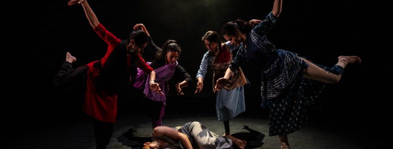 A group of four dancers pose around a fifth, who is lying on the floor as if dead or injured.