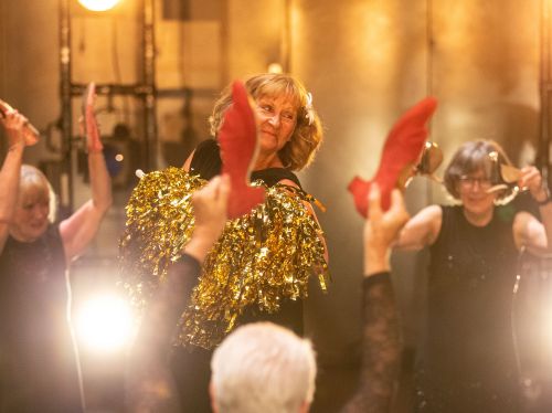 A group of performers on stage holding red props, with one person in the centre holding shiny gold tinsel-like props. Bright stage lights illuminate the scene. 